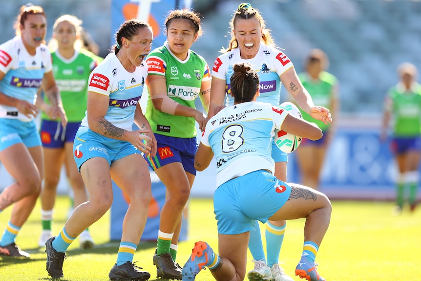 A Gold Coast Titans NRLW player on her knees after scoring a try, looks up at her excited teammates. 