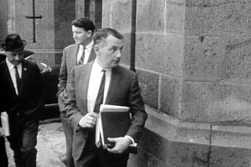 A black-and-white historical image of a lawyer carrying a stack of books walking into a stone courthouse