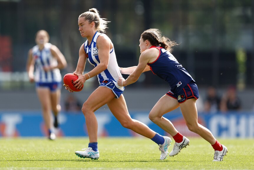 A North Melbourne AFLW player has her shirt grabbed from behind as she runs with the ball.
