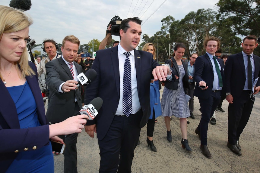 Michael Sukkar looks at his watch as journalists hold out their microphones as they pursue him