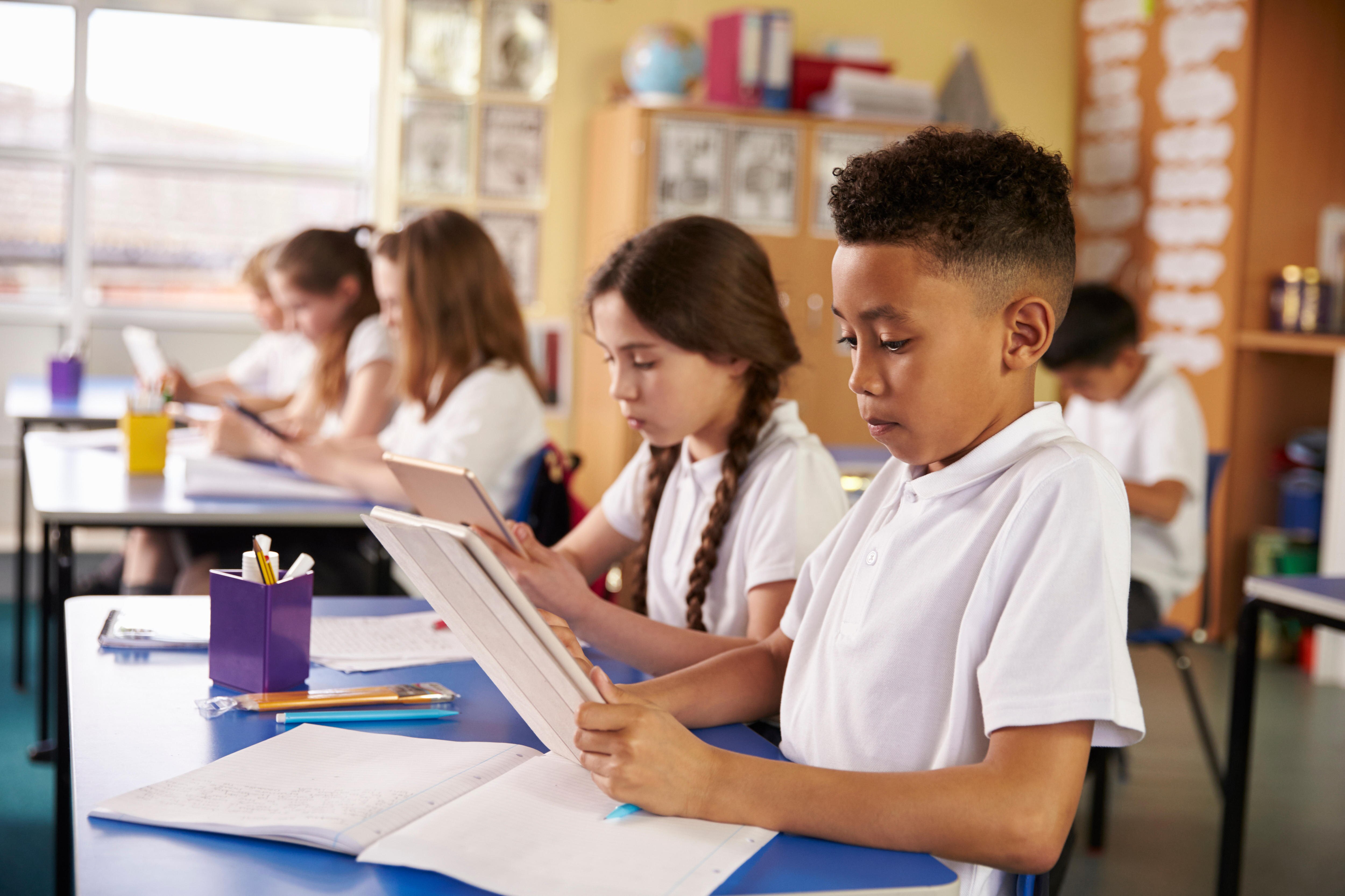 Primary school children using tablet computers while sitting at their desks in a classroom.