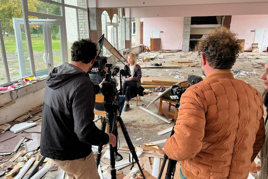 Cameraman filming a woman sitting on a chair in a room surrounded by broken glass and debris.