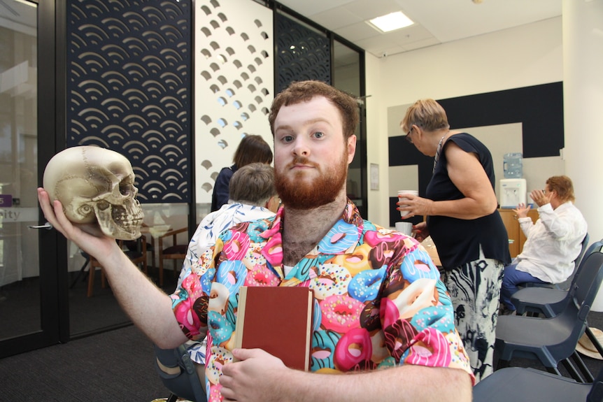 A bearded man in a donut shirt holds a skull in one hand and a book in the other.