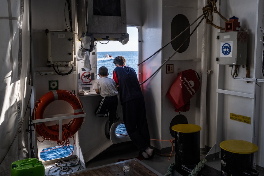 A teenager and a child look out the window from inside a ship towards the sea