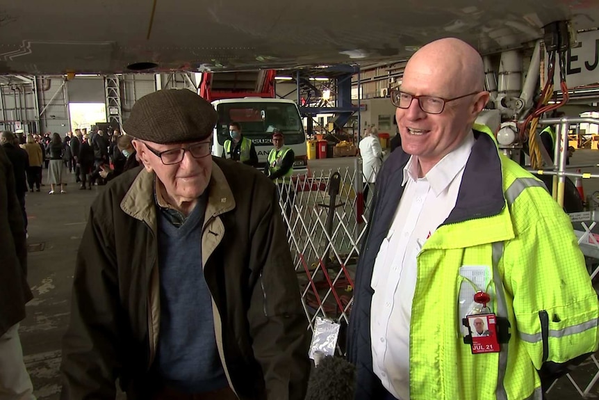 An elderly man and his son smile in an aircraft hanger