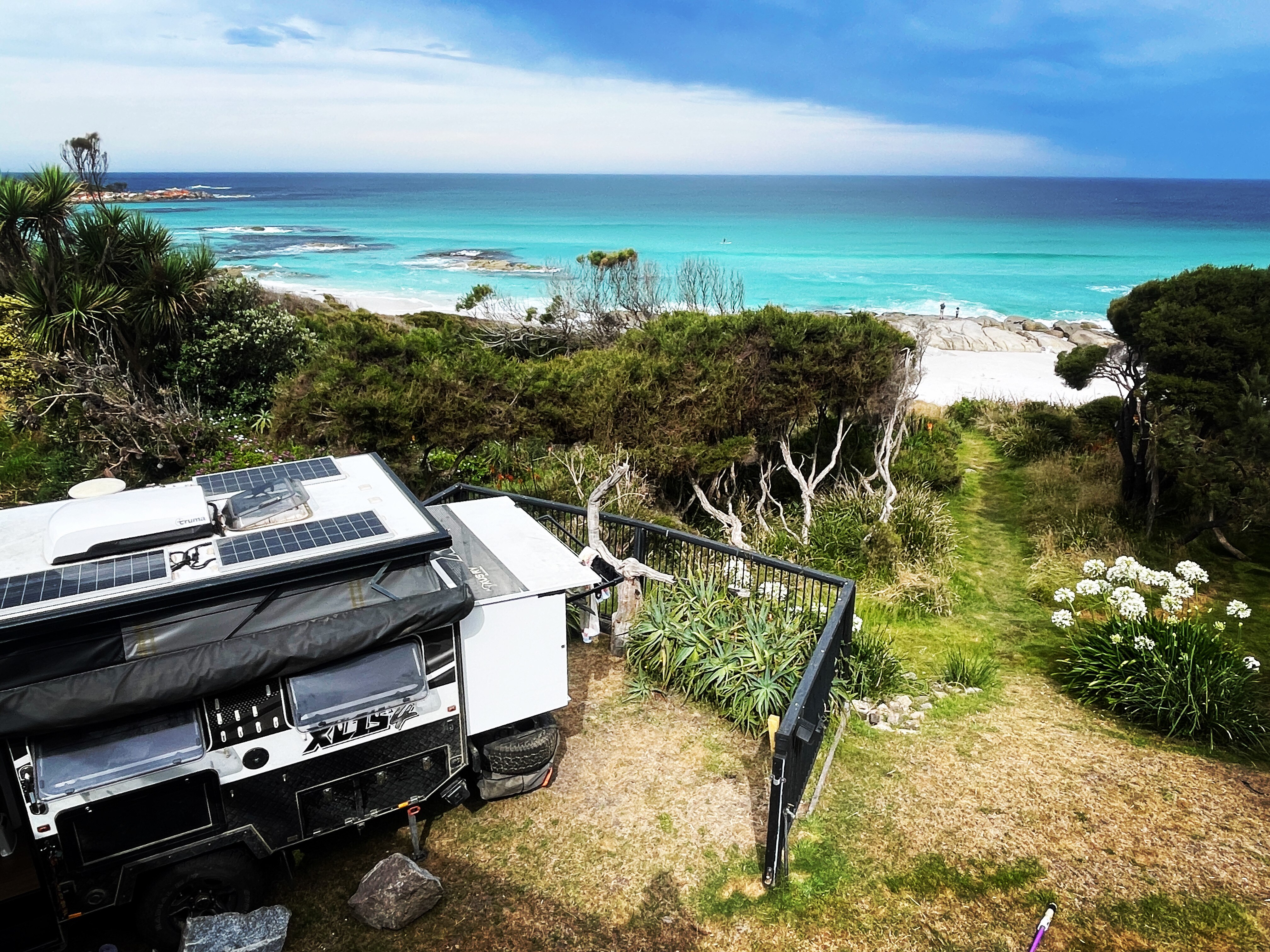 View over the roof of a caravan with 3 solar panels to a white sand beach and azure blue bay 