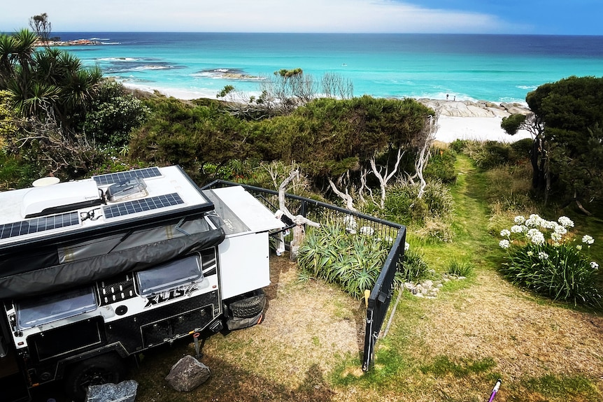 View over the roof of a caravan with 3 solar panels to a white sand beach and azure blue bay 