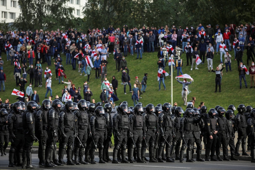 A crowd of people, many holding white-red-white flags, protest on a grassy embankment behind a row of police in black riot geat.