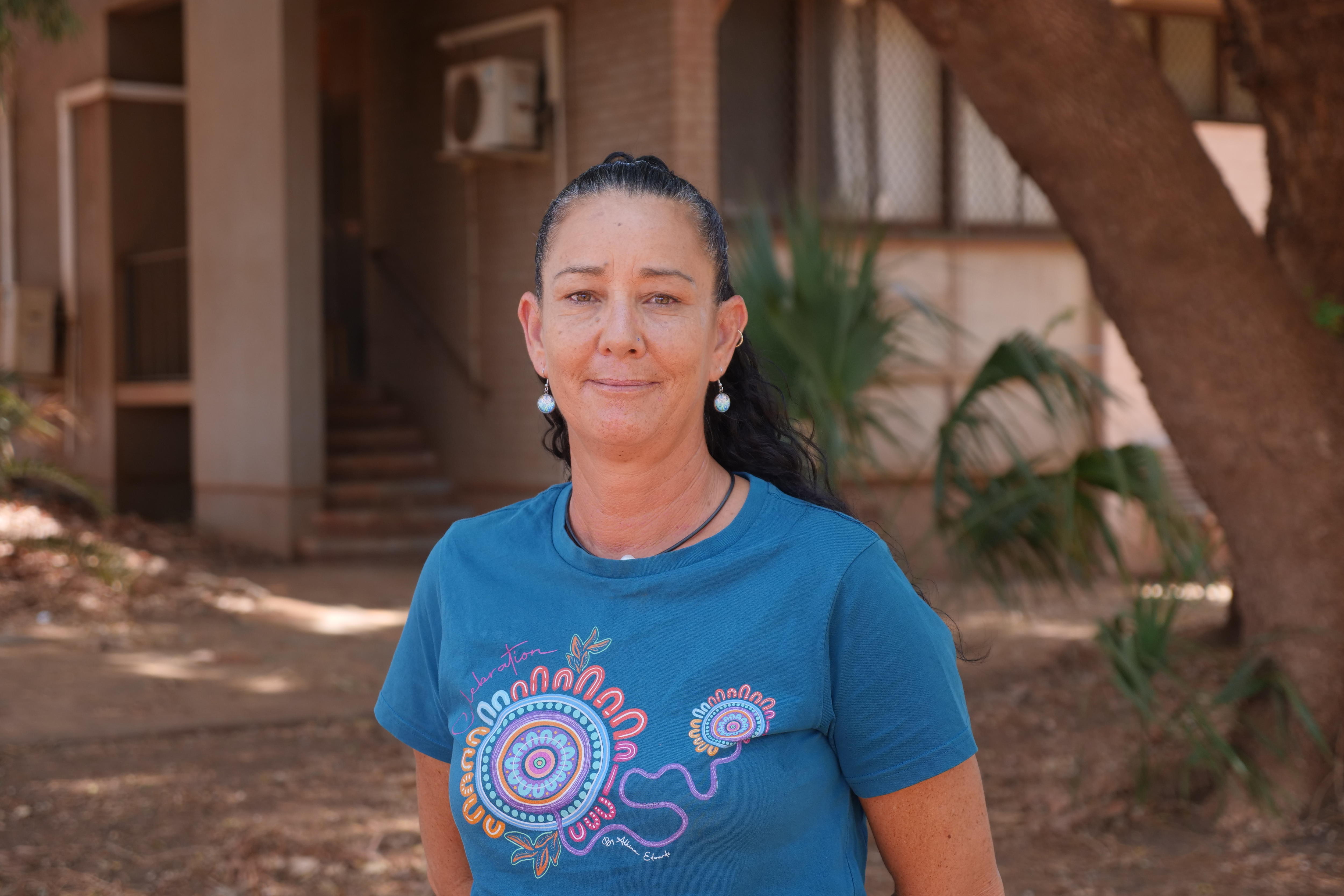 Una mujer de cabello castaño y camisa azul sonríe frente a un edificio en el exterior.