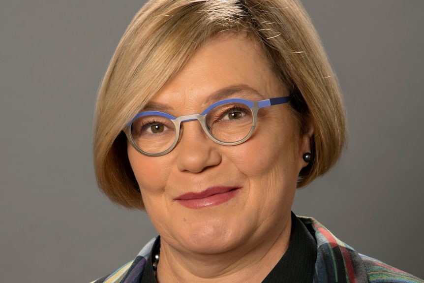A woman with a honey coloured bob and purple glasses smiles at the camera.