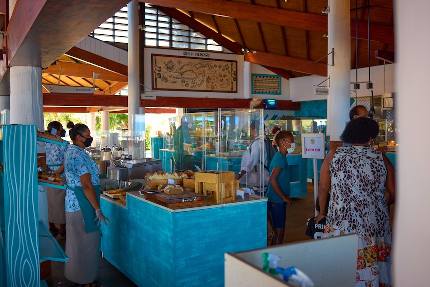 Staff in tropical uniforms and face masks stand in an open-air resort lobby behind plexiglass