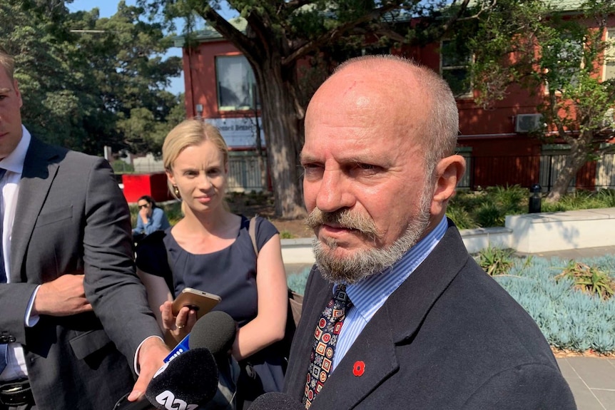 A bald man with a beard, dressed in a suit, speaks to reporters outside a court.
