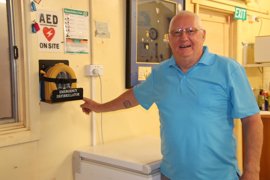 An older man wearing a blue shirt and smiling, standing next to a wall and emergency defibrillator