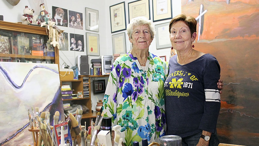 Two women stand together in front of paint brushes