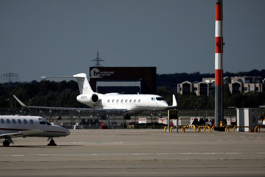 A private jet sits on tarmac at an airport, with a smaller private plane in the foreground. Buildings and powerlines in the back