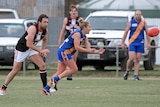 A woman footballer in a blue and gold jumper gets a handball away from a man in a red and black jumper.