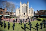 A grand church is surrounded by personell in uniform saluting a coffin entering the steps
