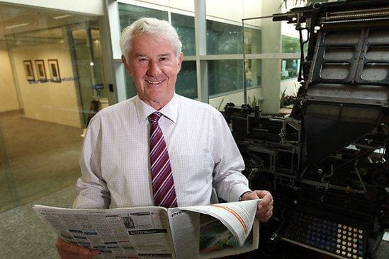 White man with grey hair, white shirt and tie holding an open newspaper in front of historic printing machine in glass foyer