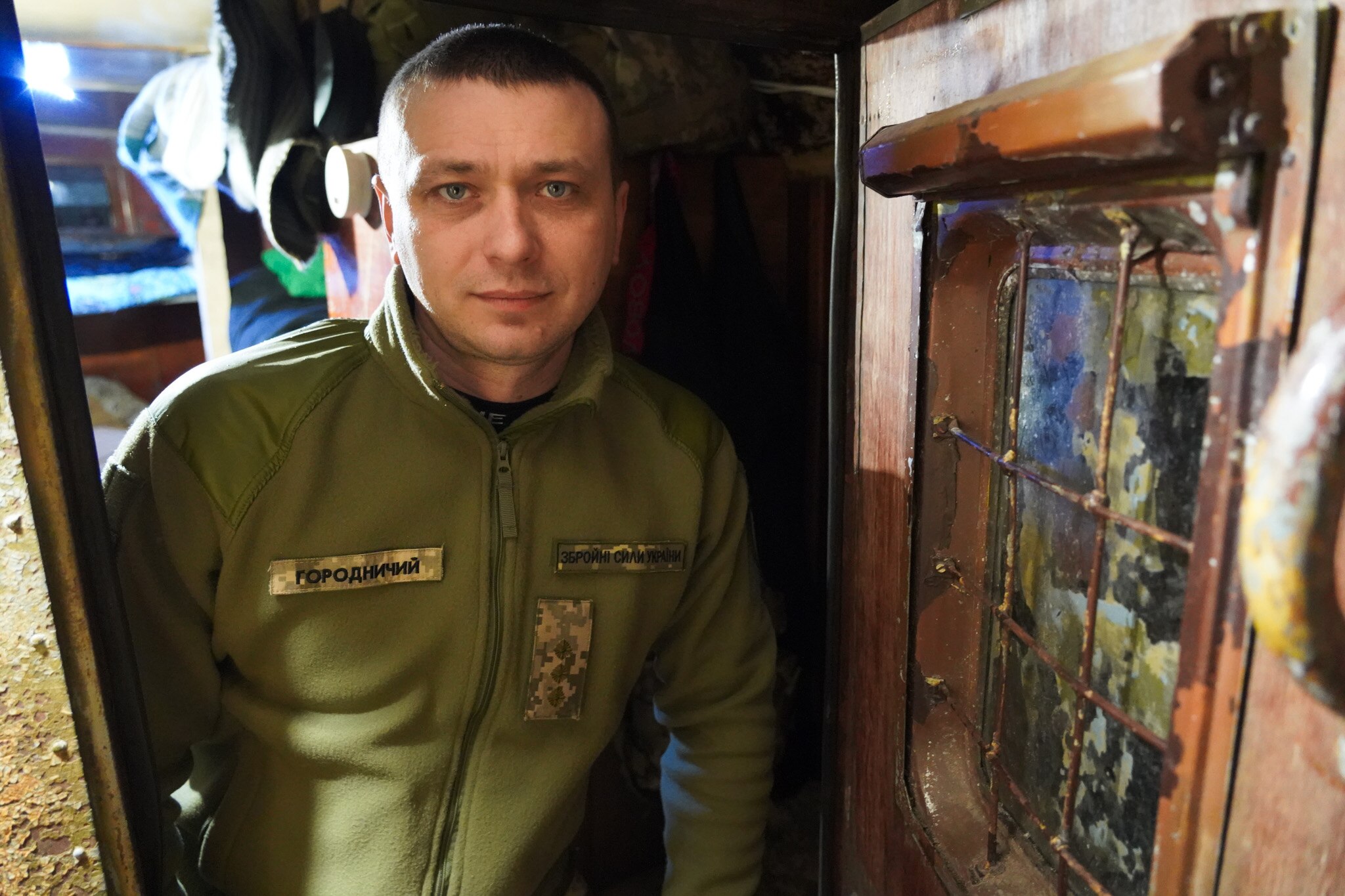 A man wearing a green jacket and badge stands in a hallway next to a rusted door.