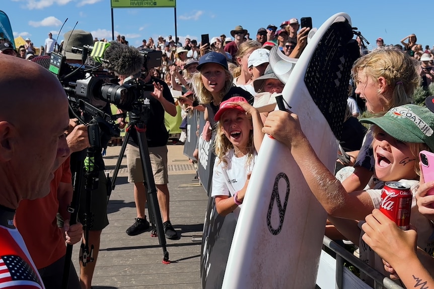 Crowds of children holding a surfboard surrounding Kelly Slater
