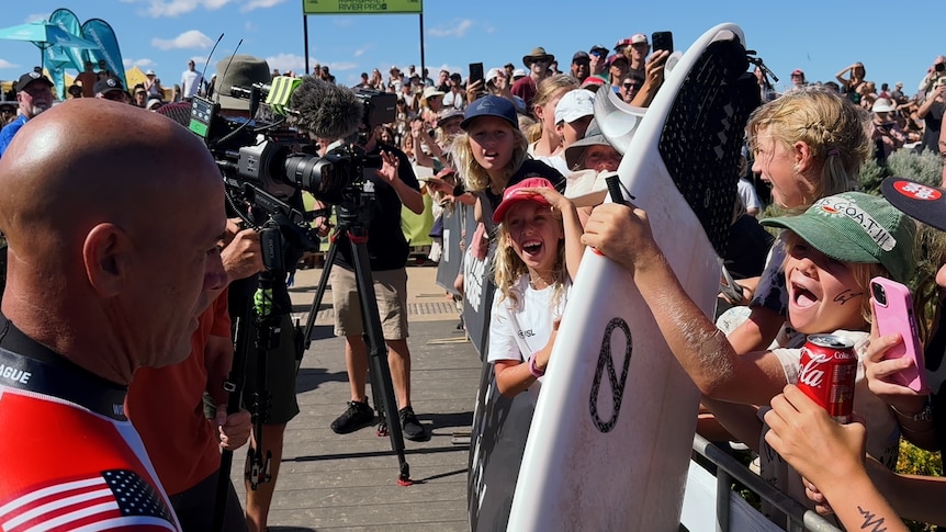 Crowds of children holding a surfboard surrounding Kelly Slater
