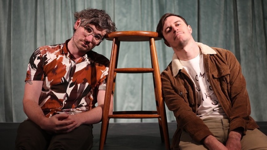 Kyran Wheatley and Alex Dyson leaning on a wooden stool in front of a green curtain on stage.