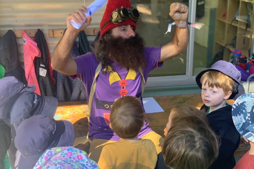 A man in a gnome costume with a huge beard looks animated in front of a group of seated small children.