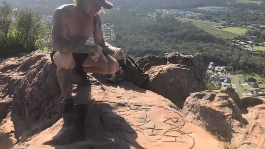 A man pointing to names in a love heart carved into rock on top of a mountain