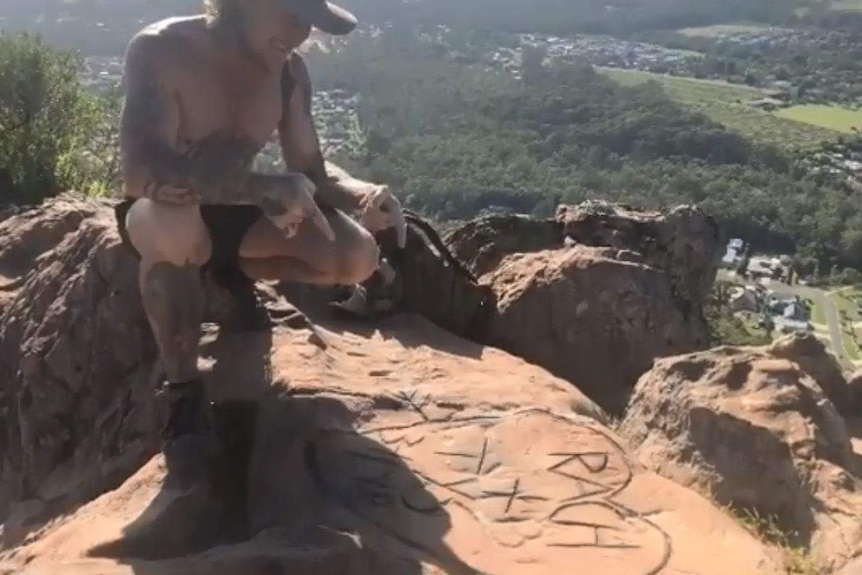 A man pointing to names in a love heart carved into rock on top of a mountain