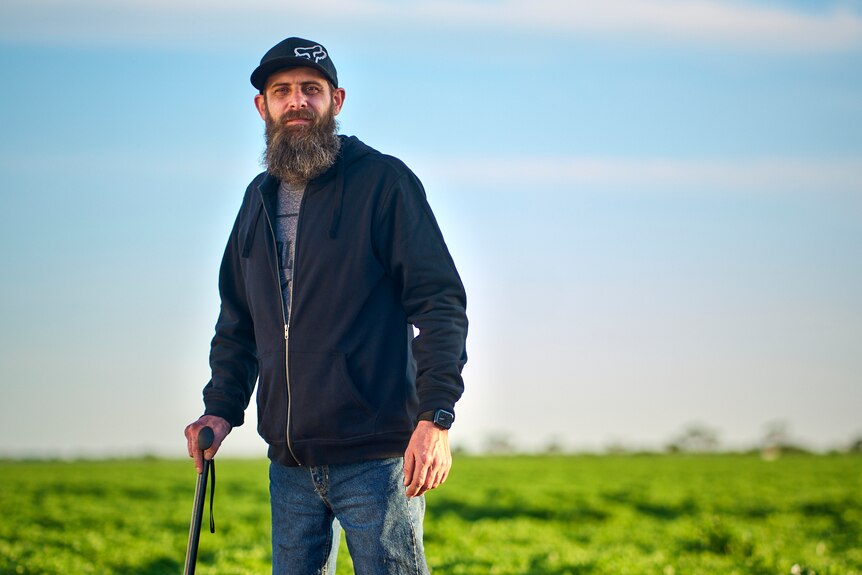 Man with two prosthetic legs leaning on a walking stick in front of a green wheat crop