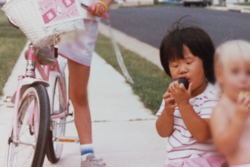 A Korean child sits down on a stool eating something as a white child perches on a bike, left, and a white baby is seen right.