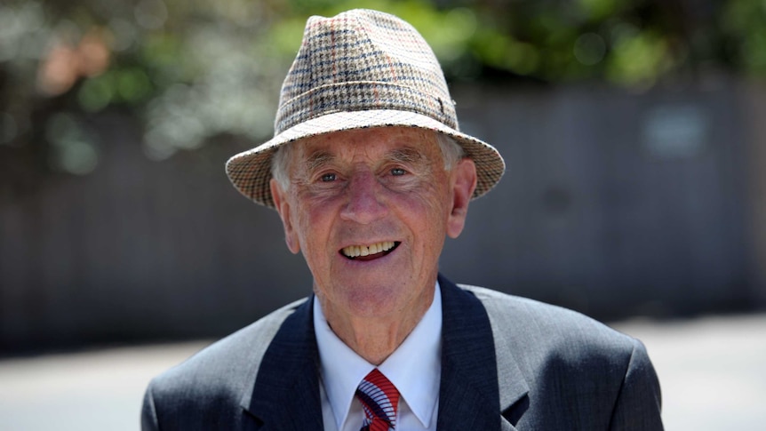 John Cain smiles as he walks along a sunny street wearing a checked hat and suit.