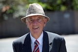 John Cain smiles as he walks along a sunny street wearing a checked hat and suit.