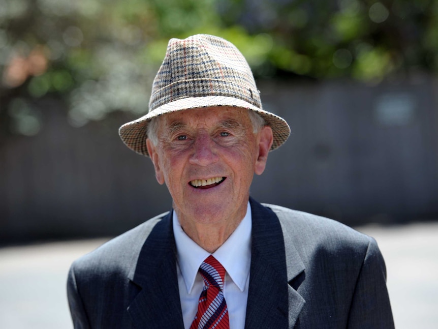 John Cain smiles as he walks along a sunny street wearing a checked hat and suit.