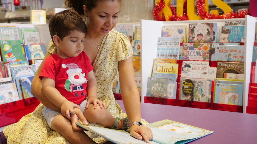 A toddler sits on his mum's lap, she is pointing to a picture in a book in front of her for him to see. 