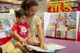 A toddler sits on his mum's lap, she is pointing to a picture in a book in front of her for him to see. 