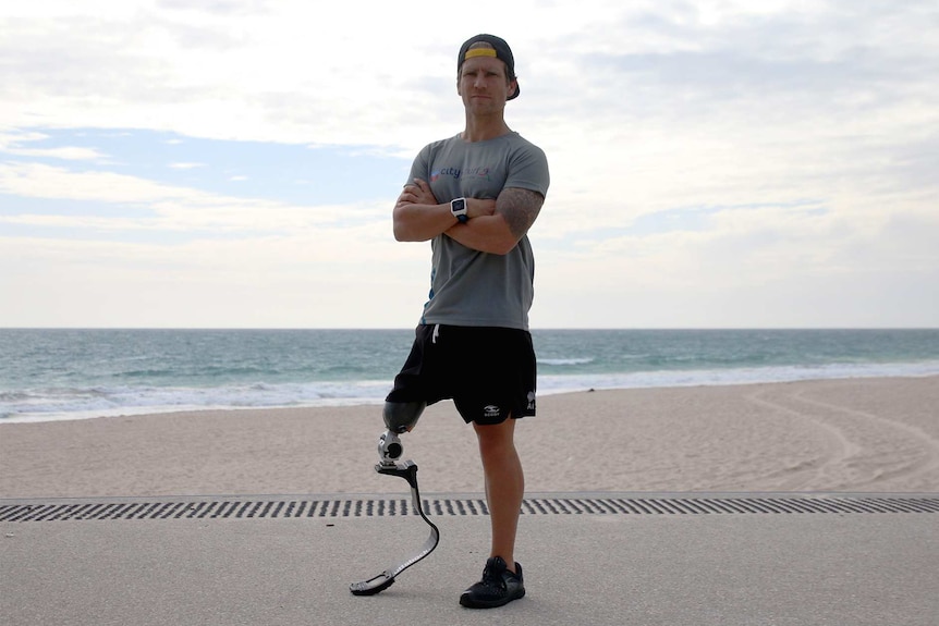 A man in a grey t-shirt and backwards black baseball cap with a prosthetic leg poses with arms crossed in front of a beach.