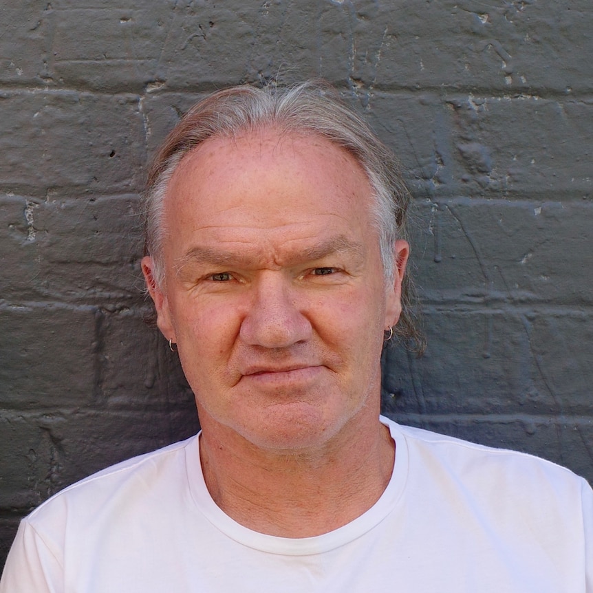 An Aboriginal man in his mid 60s with grey hair and wearing a white tshirt, stands in front of a grey wall