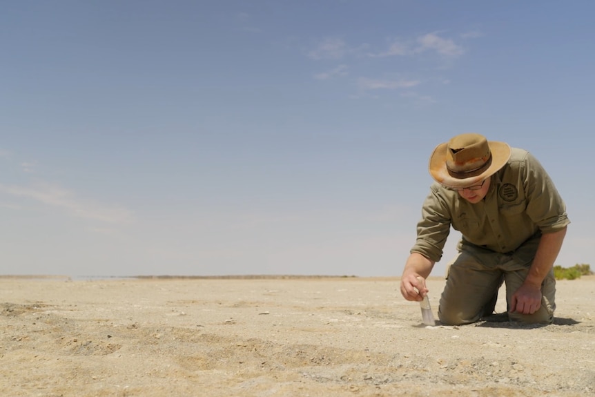 A man in khaki shirt, trousers, and hat using a paint brush to brush sand from a fossil footprint on the ground
