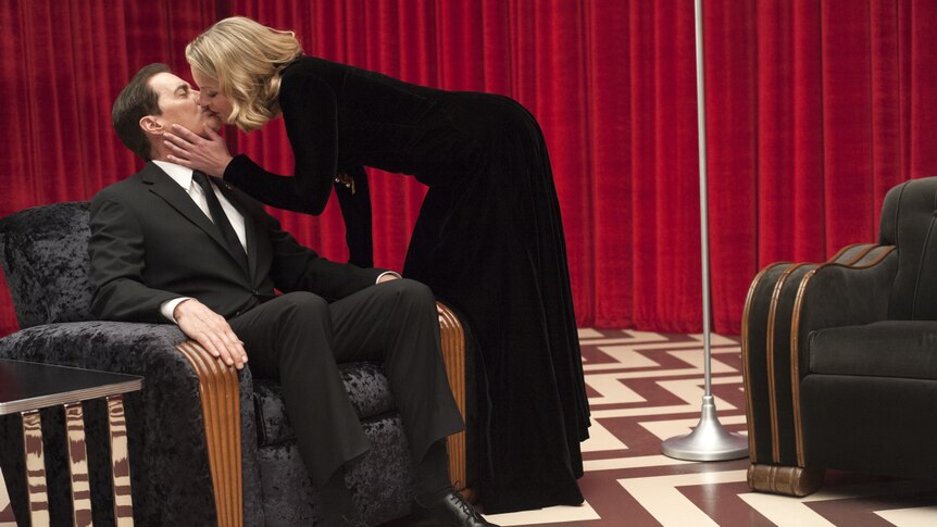A woman wearing a black dress kisses a man in a suit, sitting in a velvet chair, in a room with red curtains.
