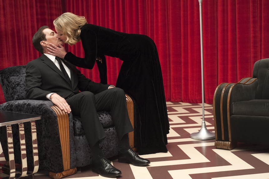 A woman wearing a black dress kisses a man in a suit, sitting in a velvet chair, in a room with red curtains.