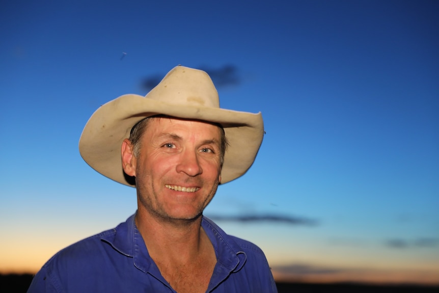 A man in a hat and blue shirt smiles at the camera as the sun sets behind him.