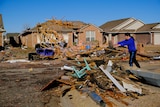 A person next to a large pile of debris with a house with a damaged roof in the background