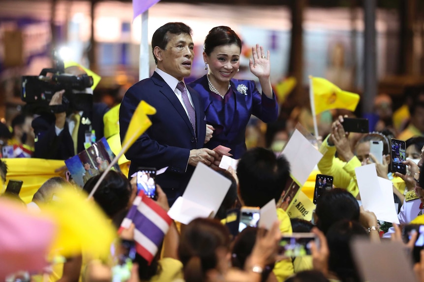 Thai King Maha Vajiralongkorn and Queen Suthida wave to supporters holding yellow flags.