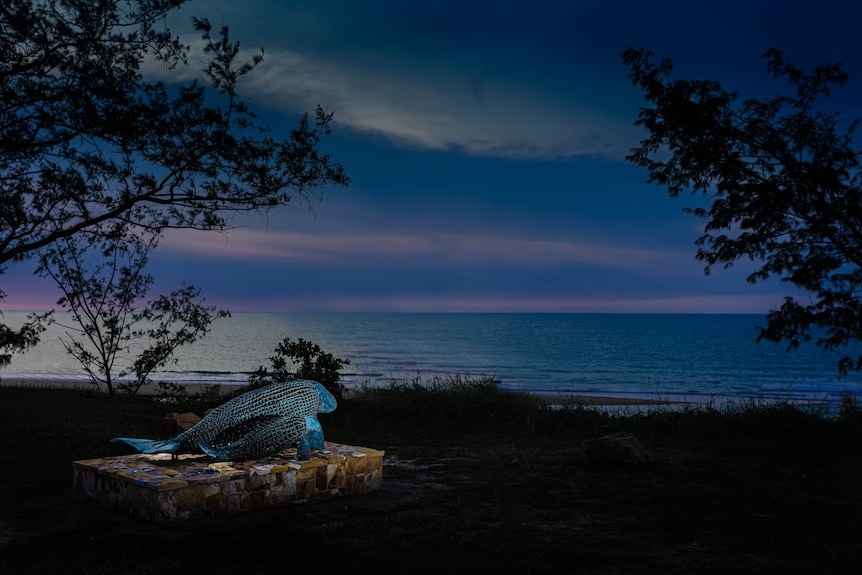 A dramatic dusk photo of a lit sculpture beside the sea.