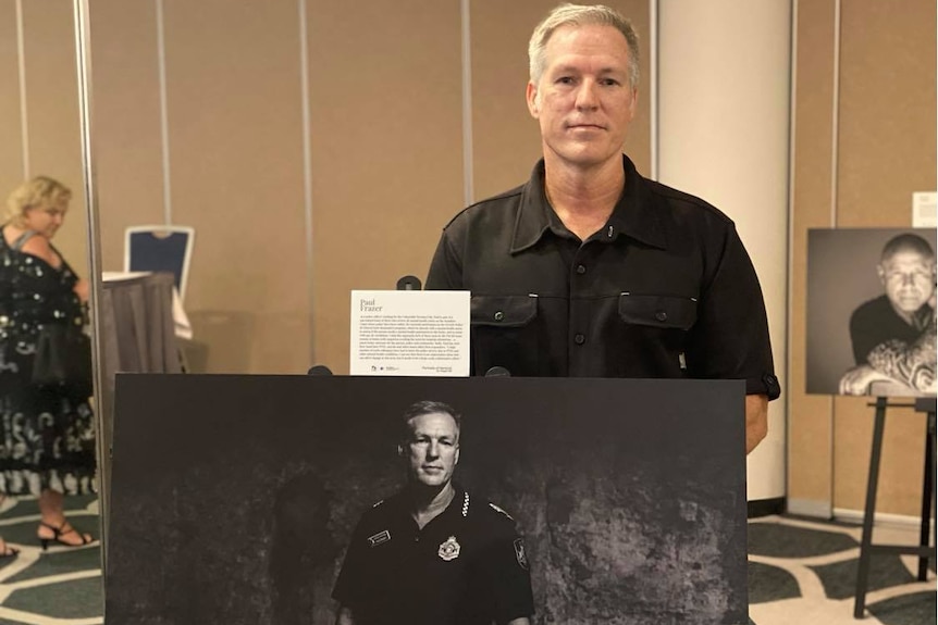 A man in a black shirt standing in front of a portrait of himself in a police shirt