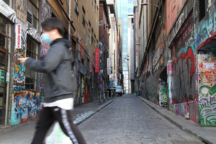 A man wearing a face mask, walking while looking at his phone in Melbourne's CBD.
