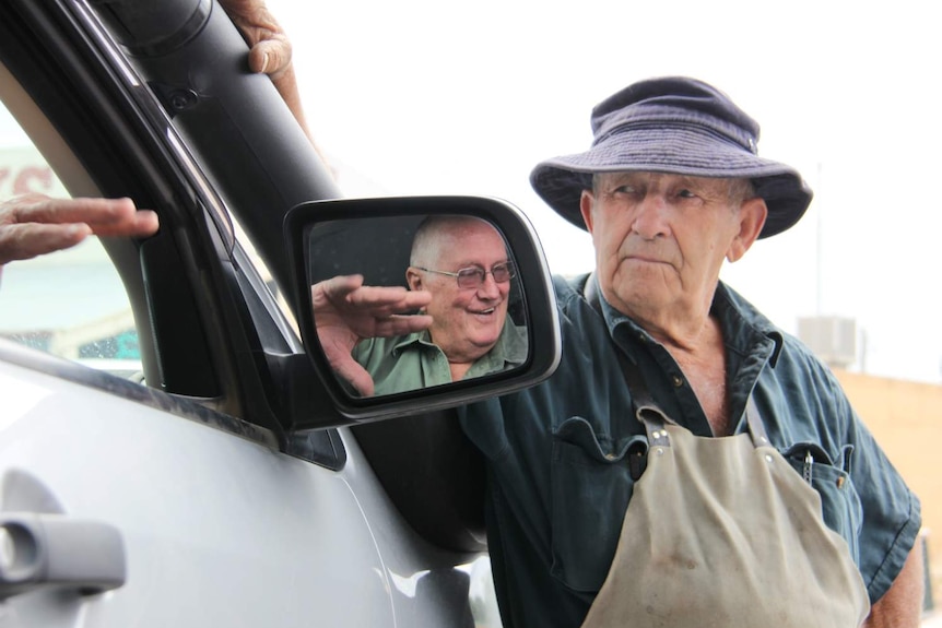 An elderly man with a floppy hat on speaks to another man, who is in a car.