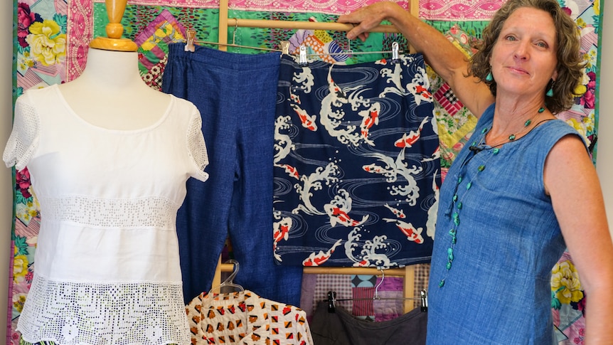 A woman, wearing a blue dress, holds up a skirt made in a sewing workshop