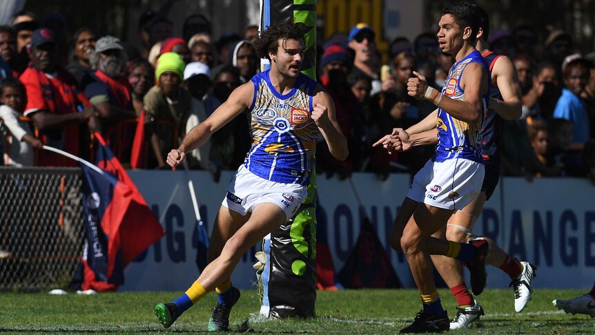 An AFL player runs away from goal in celebration after scoring, as his teammate watches him.